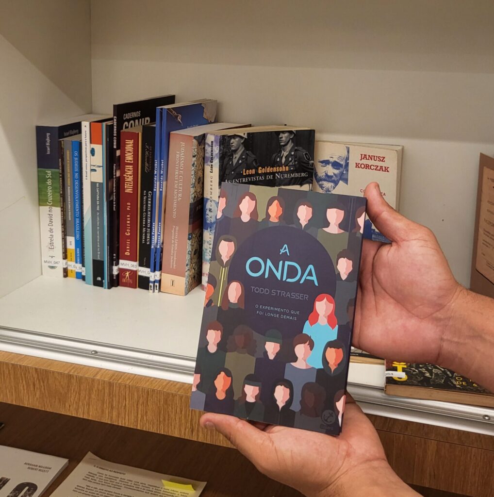 Livro "A Onda" segurado por uma mão em frente à uma estante branca com alguns livros.